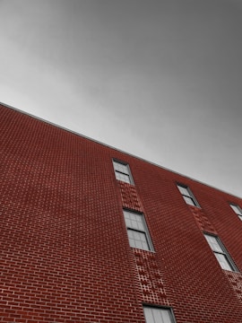 A brick building with geometric patterns in the masonry, featuring several windows. The sky is overcast and appears gray, creating a stark contrast with the rich red tones of the bricks.