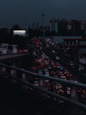 Heavy traffic fills multiple lanes of a highway under a dark, cloudy sky. The cars' lights contrast starkly against the dim atmosphere, creating a sense of congestion and movement. A billboard with the phrase 'I feel lucky' is visible on the left side.