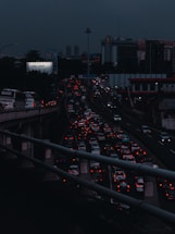 Heavy traffic fills multiple lanes of a highway under a dark, cloudy sky. The cars' lights contrast starkly against the dim atmosphere, creating a sense of congestion and movement. A billboard with the phrase 'I feel lucky' is visible on the left side.
