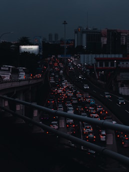 Heavy traffic fills multiple lanes of a highway under a dark, cloudy sky. The cars' lights contrast starkly against the dim atmosphere, creating a sense of congestion and movement. A billboard with the phrase 'I feel lucky' is visible on the left side.