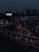 Heavy traffic fills multiple lanes of a highway under a dark, cloudy sky. The cars' lights contrast starkly against the dim atmosphere, creating a sense of congestion and movement. A billboard with the phrase 'I feel lucky' is visible on the left side.