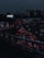 Heavy traffic fills multiple lanes of a highway under a dark, cloudy sky. The cars' lights contrast starkly against the dim atmosphere, creating a sense of congestion and movement. A billboard with the phrase 'I feel lucky' is visible on the left side.
