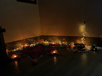 A corner with a traditional Indian decor setup featuring floor decorations with a star pattern surrounded by small oil lamps. Fairy lights are draped along the wall, creating a warm atmosphere. There are small potted plants and a plate containing some items, possibly sweets or offerings, along with marigold flowers arranged around the area.