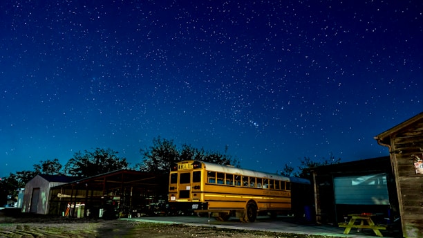 A yellow school bus is parked outside under a clear, starry night sky. The scene includes some surrounding buildings with visible structures and trees in the background. The area is illuminated by ambient light, highlighting the bus and casting subtle shadows.
