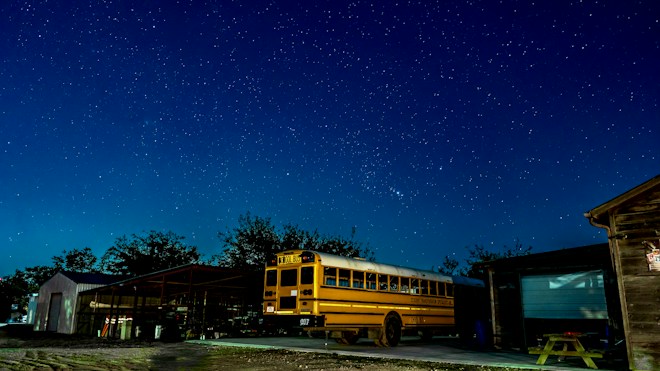 A yellow school bus is parked outside under a clear, starry night sky. The scene includes some surrounding buildings with visible structures and trees in the background. The area is illuminated by ambient light, highlighting the bus and casting subtle shadows.