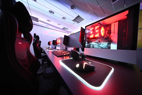 A row of high-end gaming computers on sleek desks, featuring RGB lighting and gaming accessories. The computers are housed in transparent cases with visible components, emitting a red glow. Comfortable gaming chairs with ergonomic design are positioned next to the desks, and the room's ceiling is modern with recessed lighting.