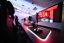A row of high-end gaming computers on sleek desks, featuring RGB lighting and gaming accessories. The computers are housed in transparent cases with visible components, emitting a red glow. Comfortable gaming chairs with ergonomic design are positioned next to the desks, and the room's ceiling is modern with recessed lighting.