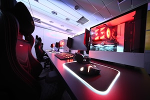 A row of high-end gaming computers on sleek desks, featuring RGB lighting and gaming accessories. The computers are housed in transparent cases with visible components, emitting a red glow. Comfortable gaming chairs with ergonomic design are positioned next to the desks, and the room's ceiling is modern with recessed lighting.