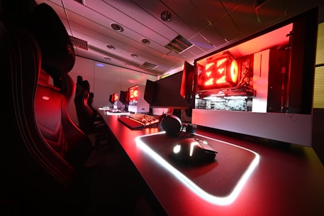 A modern gaming setup featuring several high-end computers with illuminated red interior lighting. The room is equipped with gaming chairs exhibiting a sleek black and red design. Multiple monitors are arranged on a long desk, and peripheral devices like keyboards and mice are uniformly placed, reflecting an ambiance ideal for gaming or competitive play.