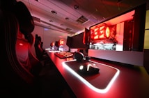 A modern gaming setup featuring several high-end computers with illuminated red interior lighting. The room is equipped with gaming chairs exhibiting a sleek black and red design. Multiple monitors are arranged on a long desk, and peripheral devices like keyboards and mice are uniformly placed, reflecting an ambiance ideal for gaming or competitive play.