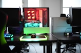 A modern gaming setup is displayed on a desk with multiple computer monitors and an open gaming PC tower. The tower is illuminated with red and green LED lights, showcasing a 'GeForce RTX' graphics card. A gaming mouse rests on a mousepad with glowing edges, and there is a keyboard with vibrant backlighting. The room appears well-lit with visible windows in the background.