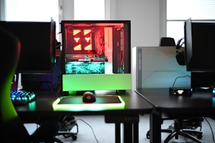 A modern gaming setup is displayed on a desk with multiple computer monitors and an open gaming PC tower. The tower is illuminated with red and green LED lights, showcasing a 'GeForce RTX' graphics card. A gaming mouse rests on a mousepad with glowing edges, and there is a keyboard with vibrant backlighting. The room appears well-lit with visible windows in the background.