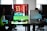 A modern gaming setup is displayed on a desk with multiple computer monitors and an open gaming PC tower. The tower is illuminated with red and green LED lights, showcasing a 'GeForce RTX' graphics card. A gaming mouse rests on a mousepad with glowing edges, and there is a keyboard with vibrant backlighting. The room appears well-lit with visible windows in the background.