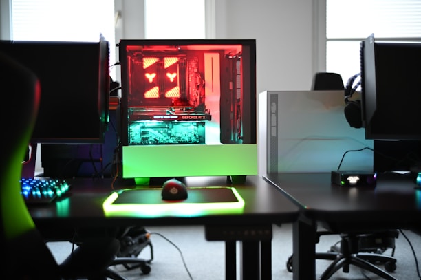 A modern gaming setup is displayed on a desk with multiple computer monitors and an open gaming PC tower. The tower is illuminated with red and green LED lights, showcasing a 'GeForce RTX' graphics card. A gaming mouse rests on a mousepad with glowing edges, and there is a keyboard with vibrant backlighting. The room appears well-lit with visible windows in the background.