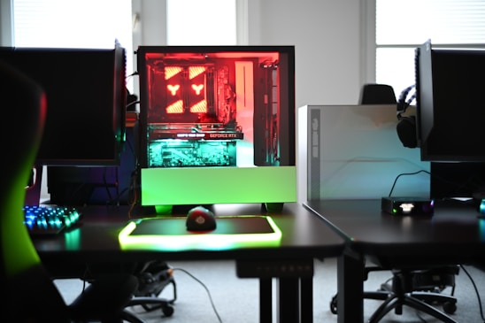 A modern gaming setup is displayed on a desk with multiple computer monitors and an open gaming PC tower. The tower is illuminated with red and green LED lights, showcasing a 'GeForce RTX' graphics card. A gaming mouse rests on a mousepad with glowing edges, and there is a keyboard with vibrant backlighting. The room appears well-lit with visible windows in the background.
