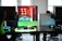A modern gaming setup is displayed on a desk with multiple computer monitors and an open gaming PC tower. The tower is illuminated with red and green LED lights, showcasing a 'GeForce RTX' graphics card. A gaming mouse rests on a mousepad with glowing edges, and there is a keyboard with vibrant backlighting. The room appears well-lit with visible windows in the background.