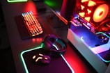 A gaming setup with a mechanical keyboard featuring multicolor RGB lighting, a glowing mouse on a mousepad with illuminated edges, a set of headphones, and a high-end gaming PC with a transparent side panel showcasing internal components and red LED lights.