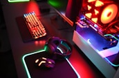 A gaming setup with a mechanical keyboard featuring multicolor RGB lighting, a glowing mouse on a mousepad with illuminated edges, a set of headphones, and a high-end gaming PC with a transparent side panel showcasing internal components and red LED lights.