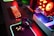 A gaming setup with a mechanical keyboard featuring multicolor RGB lighting, a glowing mouse on a mousepad with illuminated edges, a set of headphones, and a high-end gaming PC with a transparent side panel showcasing internal components and red LED lights.