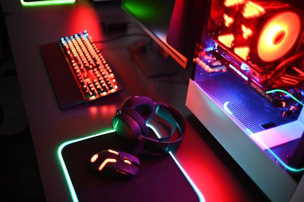 A gaming setup with a mechanical keyboard featuring multicolor RGB lighting, a glowing mouse on a mousepad with illuminated edges, a set of headphones, and a high-end gaming PC with a transparent side panel showcasing internal components and red LED lights.