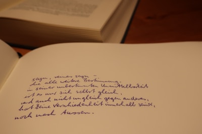 A close-up of an open notebook with handwritten text in blue ink. The writing appears to be in a poetic style. In the background, there is a partially visible closed book on a wooden surface.