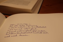 A close-up of an open notebook with handwritten text in blue ink. The writing appears to be in a poetic style. In the background, there is a partially visible closed book on a wooden surface.