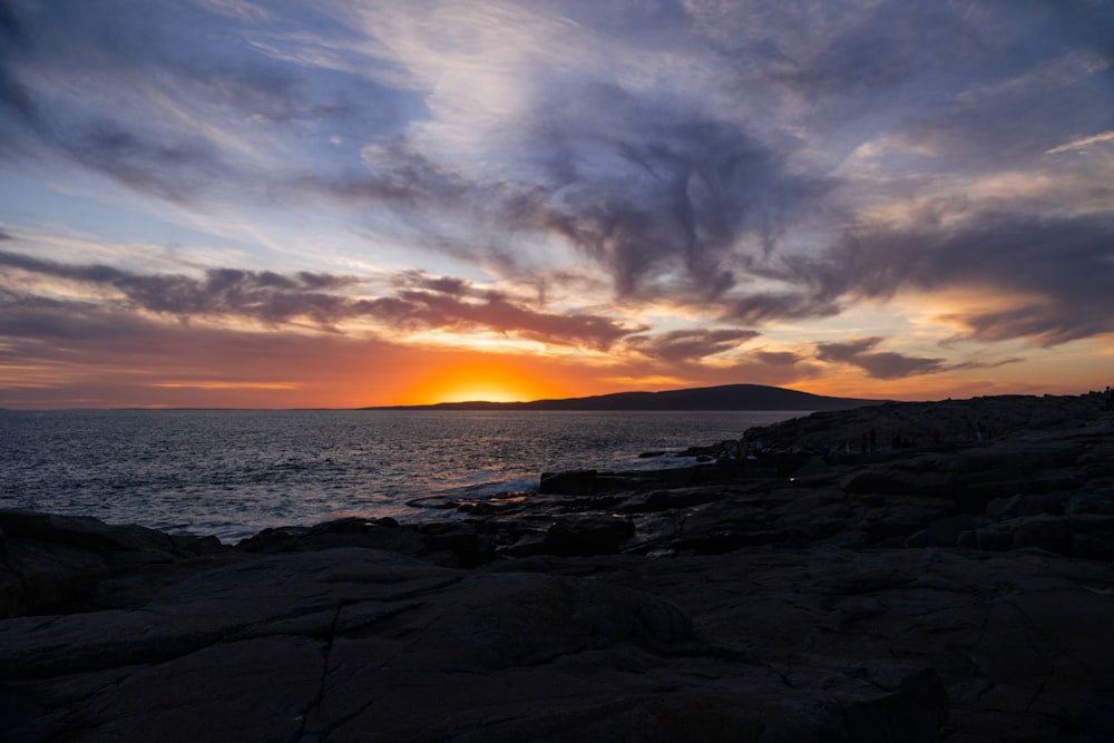 the sun is setting over the ocean with rocks in front of it