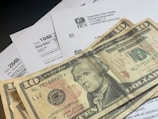 Several U.S. ten-dollar bills are placed on top of IRS tax forms and documents, including Form 1040 and envelopes addressed from the Department of the Treasury and Internal Revenue Service.