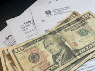 Several U.S. ten-dollar bills are placed on top of IRS tax forms and documents, including Form 1040 and envelopes addressed from the Department of the Treasury and Internal Revenue Service.