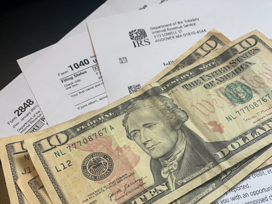 Several U.S. ten-dollar bills are placed on top of IRS tax forms and documents, including Form 1040 and envelopes addressed from the Department of the Treasury and Internal Revenue Service.