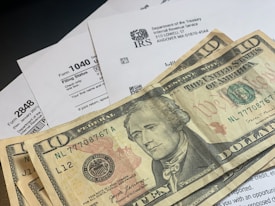 Several U.S. ten-dollar bills are placed on top of IRS tax forms and documents, including Form 1040 and envelopes addressed from the Department of the Treasury and Internal Revenue Service.