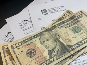 Several U.S. ten-dollar bills are placed on top of IRS tax forms and documents, including Form 1040 and envelopes addressed from the Department of the Treasury and Internal Revenue Service.