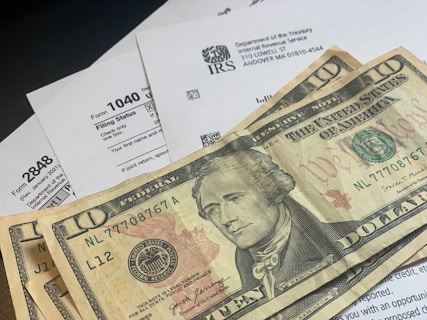 Several U.S. ten-dollar bills are placed on top of IRS tax forms and documents, including Form 1040 and envelopes addressed from the Department of the Treasury and Internal Revenue Service.