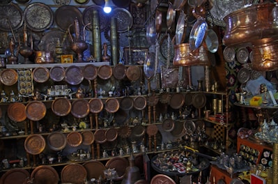 A collection of handcrafted metal items, predominantly copper and brass, displayed on shelves and hanging from the ceiling. The assortment includes trays, pots, and pitchers, featuring intricate designs and patterns. The setting has a warm, vintage feel, with ambient lighting enhancing the metallic sheen.