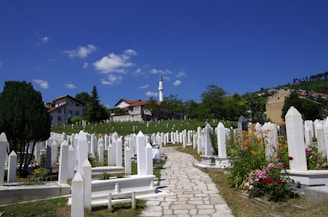 A serene cemetery scene with white tombstones lining a stone path. The graves are surrounded by vibrant flowers and greenery, set against a backdrop of houses and a minaret rising in the distance. The sky is clear with a few fluffy white clouds, adding to the peaceful atmosphere.