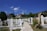 A serene cemetery scene with white tombstones lining a stone path. The graves are surrounded by vibrant flowers and greenery, set against a backdrop of houses and a minaret rising in the distance. The sky is clear with a few fluffy white clouds, adding to the peaceful atmosphere.