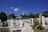 A serene cemetery scene with white tombstones lining a stone path. The graves are surrounded by vibrant flowers and greenery, set against a backdrop of houses and a minaret rising in the distance. The sky is clear with a few fluffy white clouds, adding to the peaceful atmosphere.