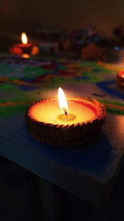 A close-up view of a lit clay diya with a bright flame, placed on a colored surface. The background has additional unfocused diyas, and a mixture of vibrant colors can be seen providing a festive atmosphere.