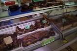 A display of various cuts of cured meat in a butcher shop, including different sizes and shapes of dark brown and reddish-brown meats, some of which are labeled with prices. The meats are placed inside a refrigerated glass case and there are trays and tools visible in the background.