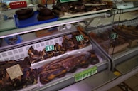 A display of various cuts of cured meat in a butcher shop, including different sizes and shapes of dark brown and reddish-brown meats, some of which are labeled with prices. The meats are placed inside a refrigerated glass case and there are trays and tools visible in the background.