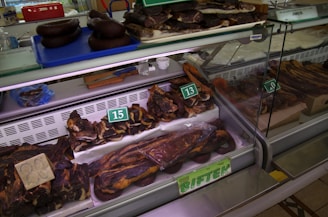 A display of various cuts of cured meat in a butcher shop, including different sizes and shapes of dark brown and reddish-brown meats, some of which are labeled with prices. The meats are placed inside a refrigerated glass case and there are trays and tools visible in the background.
