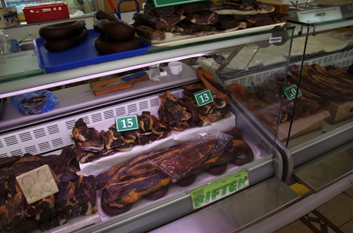 A display of various cuts of cured meat in a butcher shop, including different sizes and shapes of dark brown and reddish-brown meats, some of which are labeled with prices. The meats are placed inside a refrigerated glass case and there are trays and tools visible in the background.