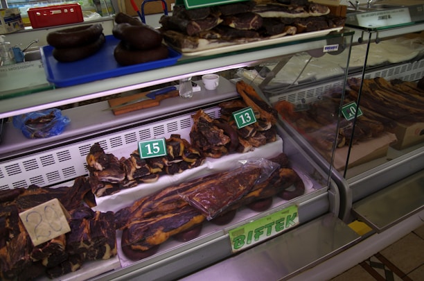 A display of various cuts of cured meat in a butcher shop, including different sizes and shapes of dark brown and reddish-brown meats, some of which are labeled with prices. The meats are placed inside a refrigerated glass case and there are trays and tools visible in the background.