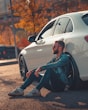 a man sitting on the ground next to a white car