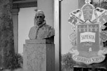 A grayscale image featuring a stone bust of a figure with spectacles, positioned on a pedestal. This bust is situated outdoors with some architectural elements visible in the background. To the right, there's a reflective surface displaying a coat of arms or emblem bearing the word 'SAPENTIA.' The scene conveys a historical or academic setting.