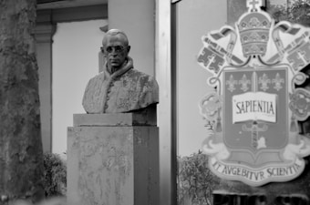 A grayscale image featuring a stone bust of a figure with spectacles, positioned on a pedestal. This bust is situated outdoors with some architectural elements visible in the background. To the right, there's a reflective surface displaying a coat of arms or emblem bearing the word 'SAPENTIA.' The scene conveys a historical or academic setting.