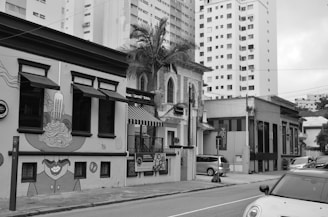 A city street scene featuring a row of distinct buildings, one of which is decorated with artistic murals and signage advertising pasta, cannoli, and drinks. The architecture includes a mix of modern and older styles with tall residential buildings in the background. Vehicles are parked along the street, and there are palm trees visible against the buildings.