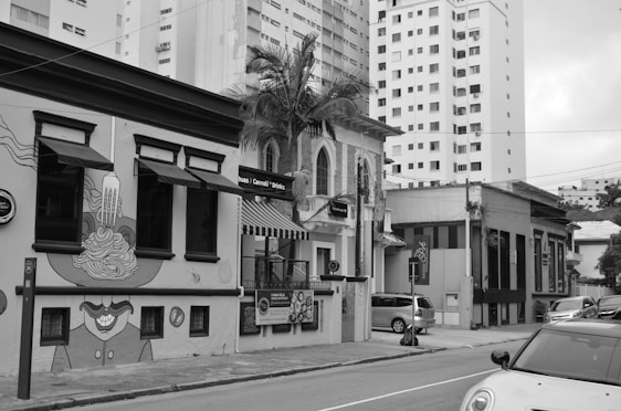 A city street scene featuring a row of distinct buildings, one of which is decorated with artistic murals and signage advertising pasta, cannoli, and drinks. The architecture includes a mix of modern and older styles with tall residential buildings in the background. Vehicles are parked along the street, and there are palm trees visible against the buildings.