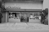 A building facade with a modern design, featuring a sign labeled 'a.Oculista'. The store has large windows displaying various items inside. Potted plants are positioned on both sides of the entrance. The exterior is characterized by a perforated pattern above the windows, and there appears to be a seating area with a person inside.