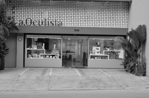 A building facade with a modern design, featuring a sign labeled 'a.Oculista'. The store has large windows displaying various items inside. Potted plants are positioned on both sides of the entrance. The exterior is characterized by a perforated pattern above the windows, and there appears to be a seating area with a person inside.